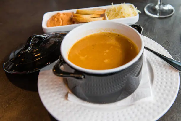 Studio photo of a small pot with lobster and fish soup. Rouille, bread and cheese are served on a side plate.