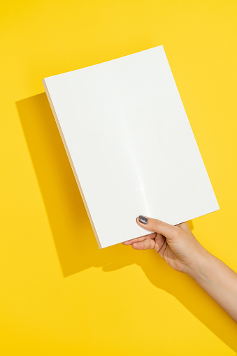 Hand holding blank white cover magazine mockup, template on yellow background