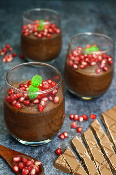 istock Image of three glasses containing home-made chocolate mousse dessert topped with pomegranate seeds and mint leaf garnish beside milk chocolate bar chunks and wooden spoon of pomegranate seeds, dark grey background, focus on foreground 1321778877