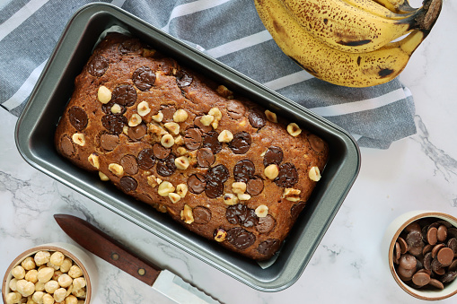 Stock photo showing elevated view of cake loaf tin containing homemade banana loaf, besides a hand of bananas, bowls of chocolate chips and hazel nuts, a tea towel and knife, on a marble effect background.
