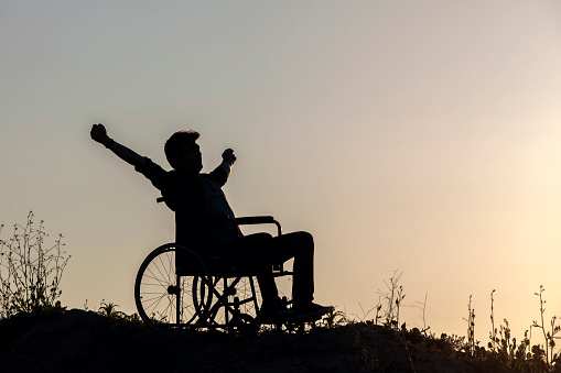 Never give up. Rear view of young handicapped man sitting on wheelchair on top of mountain with hands up and looking at amazing nature landscape while traveling alone. International Disability Day