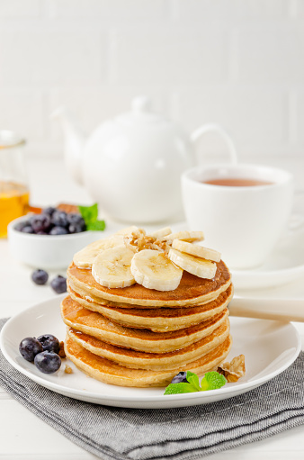 A stack of oatmeal banana pancakes with slices of fresh bananas, walnuts and honey on top with cup of tea on a white wooden background. A healthy breakfast. Copy space