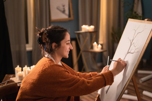 an art student sits in her home living room in the evening and draws a sketch of a tree, nature for a college credit. the passionate woman smiles, satisfied with painting - drawing sketch artist charcoal drawing imagens e fotografias de stock