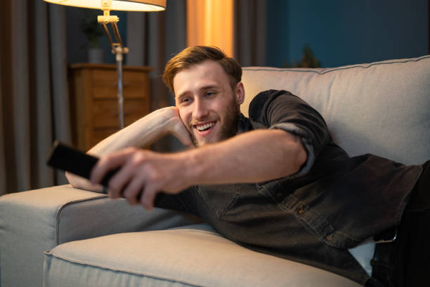 istock Laughing, smiling young man, resting on sofa in living room, lying on couch lengthwise, supporting himself with arm under head, holding elbow on armrest of sofa, holding TV remote, turning on cabaret 1321854618