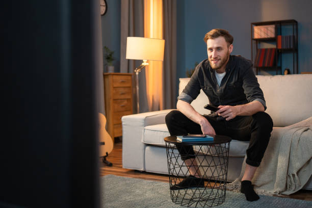 istock Happy student resting on sofa in the living room, next to him there is a coffee table on which there is a book and a phone, a boy is sitting in front of the TV changing channels with a remote control 1321854640