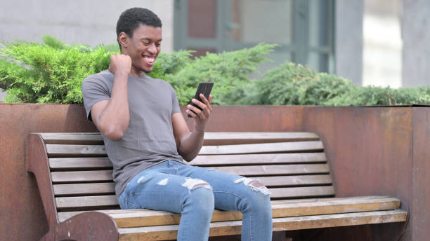 ambitious young african man celebrating success on smartphone - using phone garden bench imagens e fotografias de stock