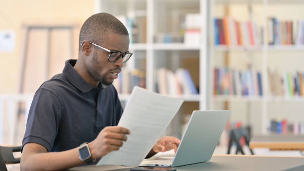 istock Young African Man with Laptop doing Paperwork in Library 1321867747