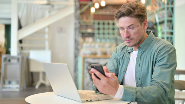 istock Smartphone use by Middle Aged Man with Laptop in Cafe 1321870774