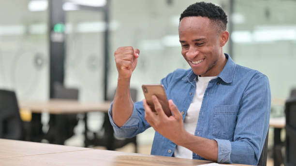 istock Casual African Man Celebrating Success on Smartphone in Office 1321871987