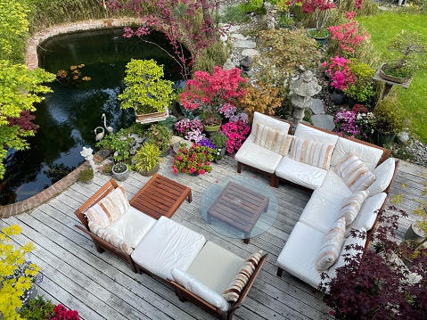 Image of outdoor lounging area on sunny summer garden decking, grooved, whitewashed wooden deck, hardwood seating with cushions, glass table top, koi carp fish pond, bonsai trees, Japanese maples, landscaped oriental design garden, elevated view