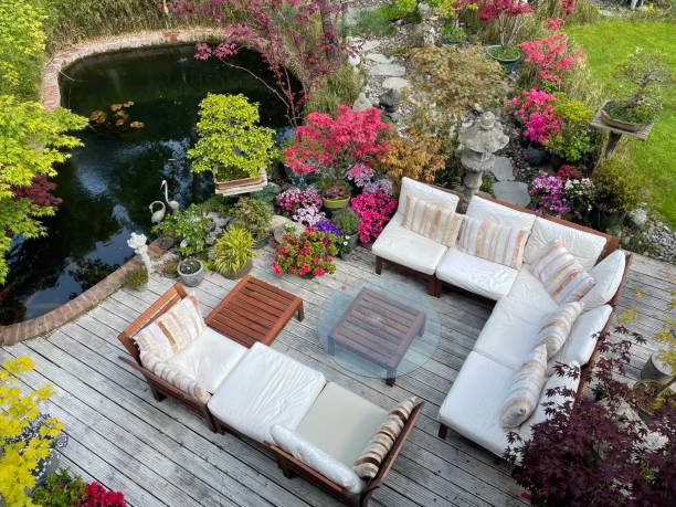 Image of outdoor lounging area on sunny summer garden decking, grooved, whitewashed wooden deck, hardwood seating with cushions, glass table top, koi carp fish pond, bonsai trees, Japanese maples, landscaped oriental design garden, elevated view Stock photo showing ornamental Japanese-style garden with outdoor lounge area. Featuring crystal clear koi pond, whitewashed, grooved timber decking patio, Japanese bonsai maples and hardwood, cushion covered seating. garden feature stock pictures, royalty-free photos & images
