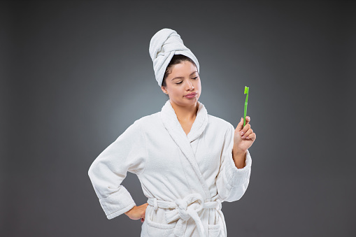 Reluctant brushing of teeth. Unhappy woman in a bathrobe and a towel wrapped around head holds a toothbrush in hand as she stands in front of a gray background and ponders whether to brush her teeth
