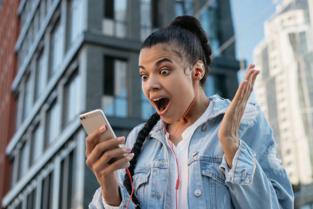 興奮したアフリカ系アメリカ人女性は、携帯電話を使用して、オンラインショッピング、デジタル画面を見て。感情的な顔を持つヒップスターの女の子、ニュース速報を読んで開いた口 - よい知らせ ストックフォトと画像