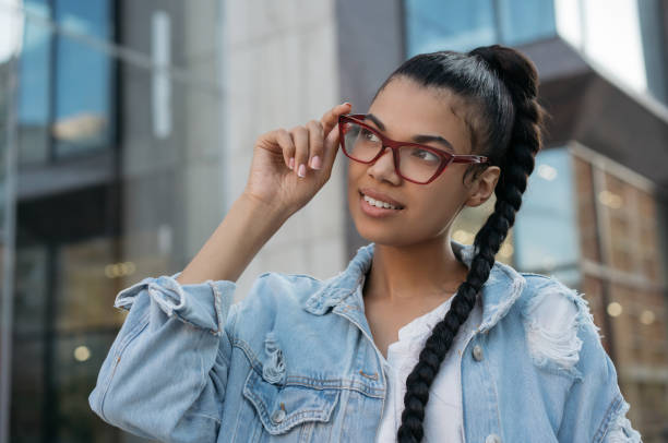 Beautiful African American woman wearing stylish eyeglasses, standing on the street and smiling. Young happy fashion model posing for pictures outdoors. Natural beauty concept stock photo