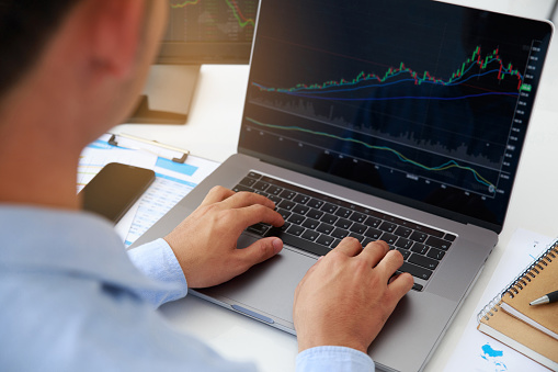 Close-up young businessman statistic graph of stock market financial indices analysis on a laptop and on a desktop computer in office. Analyzing and Trading Stock concept. Depicts TradingView financial market chart.