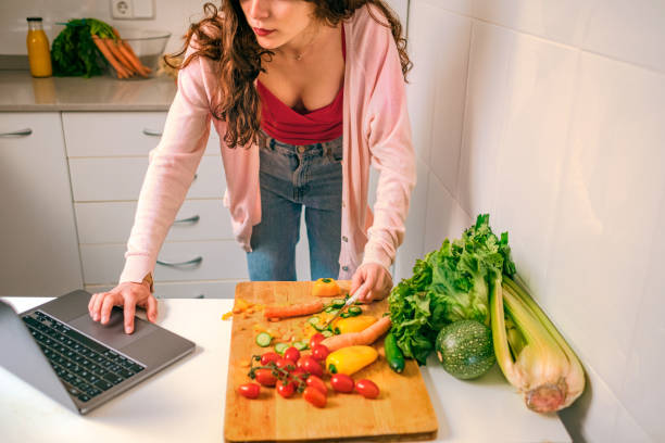 istock Young woman reading online recipes using laptop while cutting vegetables in the kitchen 1321901260