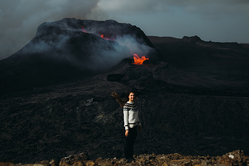 Young woman in traditional Icelandic wool sweater exploring the dramatic landscape with the new erupting volcano with the flame melting lava