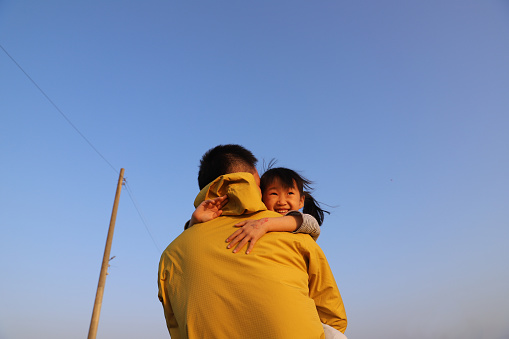 a smiling Asian girl sitting on her father's shoulder under blue sky. background for father's day