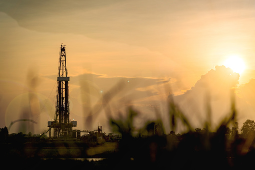 Crude oil drilling rig in silhouette with dramatic sky as background and shadow of grass peak as foreground. Petroleum exploration industrial. High contrast ratio, len flare applied and noisy photo.