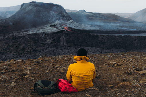 Young man backpacker in yellow jacket exploring the dramatic landscape, sitting near the big erupting volcano with melting lava in Iceland
