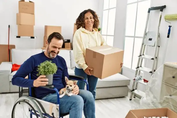 Middle age hispanic couple smiling happy. Man sitting on wheelchair with dog on his legs and woman holding cardboard box at new home.