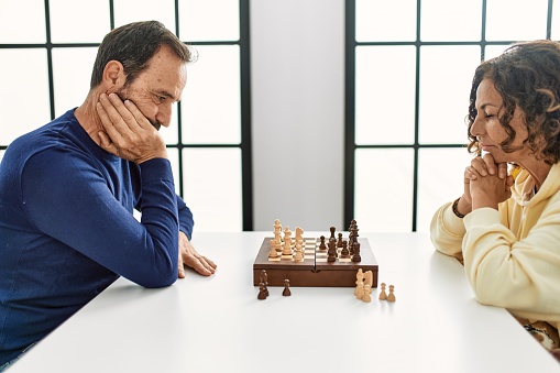 Middle age hispanic couple concentrated sitting on the table playing chess at home.