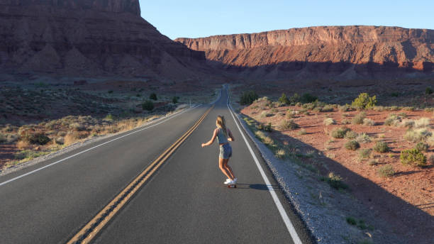 Young woman skateboards down rural road at sunrise Desert landscape and mountains surrounding single yellow line sunlight usa utah stock pictures, royalty-free photos & images