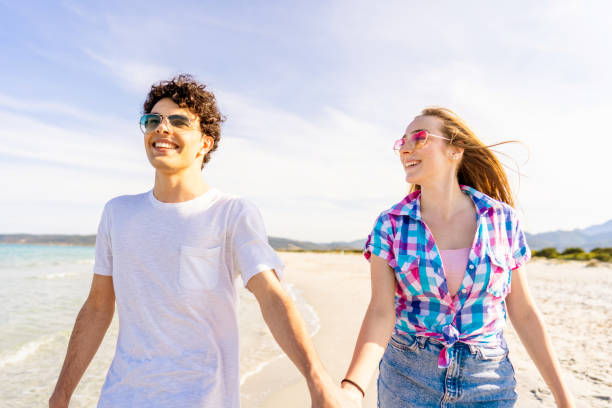 concepto de juventud despreocupada: joven muy feliz generación z pareja real cogido de la mano caminando tiernamente en la orilla del mar de la playa tropical en verano con gafas de sol. chica enamorada mira atentamente a su novioconcepto juvenil sin preo - honeymoon beach couple heterosexual couple fotografías e imágenes de stock