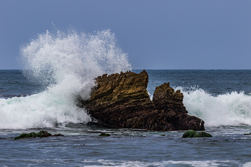 Ocean wave breaking on rock just offshore in Laguna Beach, California. White spray in the air;  blue sky in background.