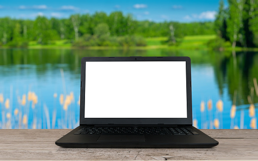 open laptop with blank screen on wooden table and summer lake or river blurred background, remote or distant work place, travel or vacation working concept