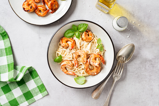 Pasta spaghetti with grilled shrimp and creamy alfredo sauce served with fresh basil leaves. Light gray background, top view
