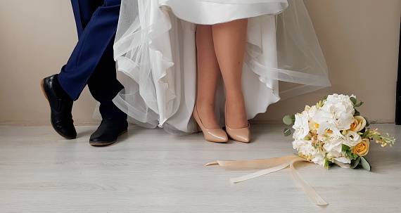 women's stiletto shoes on the bride's foot and the groom's feet in boots next to the bouquet on the floor, conceptual wedding photo
