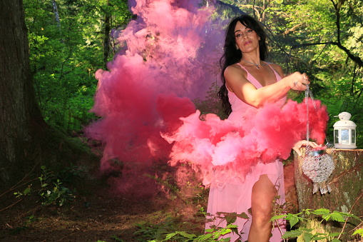 A beautiful woman with a smoke grenade and wearing a pink sleeveless dress in the forest.