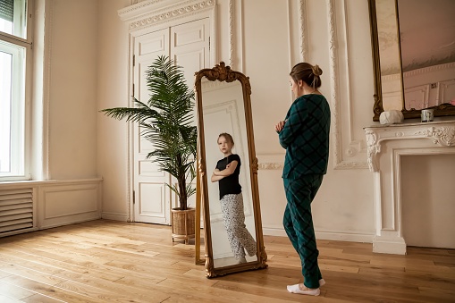 Middle age woman looking at mirror in living room and sees her daughter in reflection of teenage girl. Lonely female stands and looks in mirror in home clothes. Concept of adult life and childhood