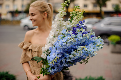 view on smiling attractive blonde woman who neatly holds in her hands large bunch of multicolored delphinium flowers