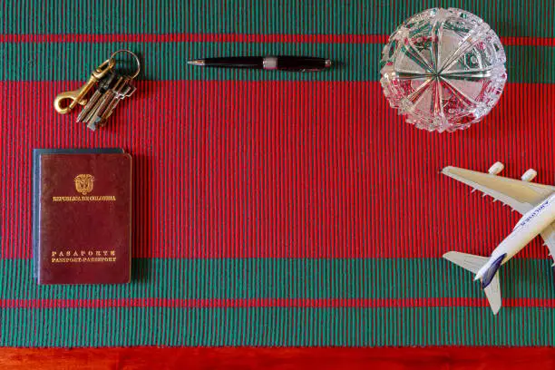 A Travel Flat Lay with a Colombia passport displayed on a red and green woven fabric which hints at Christmas. The lettering and the emblem of the Country are embossed on the cover in gold colour. In the upper half of the image is a ball-pen and to the right an airplane model. A crystal ashtray adds some pizzaz. A bunch of keys indicates a departure from home. Image shot with long exposure. Copy space.