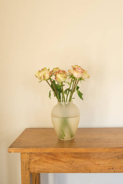 istock Cream roses in glass vase on oak side table against beige wall 1322038413