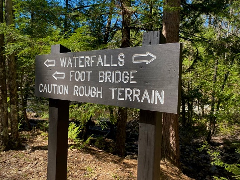 Hiking trail sign at Dave’s Falls County park in Amberg, Wisconsin. Moss covered rocks, pine trees, and sunlight showing beyond the hiking trail sign in the woods. The brown wooden sign has arrows pointing where to go to see the waterfalls and a caution “rough terrain” warning.