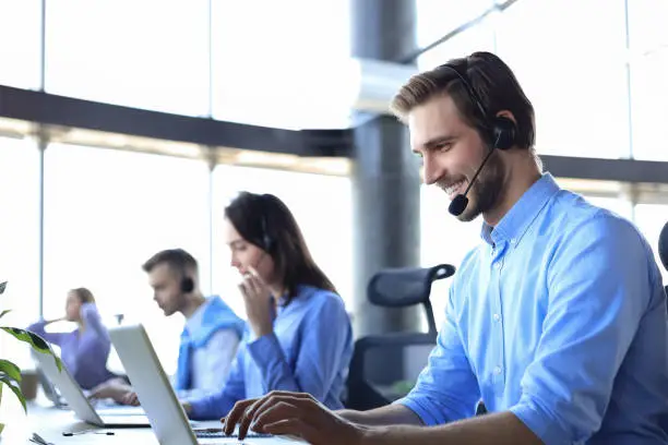 Smiling male call-center operator with headphones sitting at modern office with collegues on the backgroung, consulting online