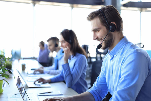 sorridente operatore di call-center maschile con cuffie sedute in un ufficio moderno con collegues sul backgroung, consulenza online. - answering foto e immagini stock