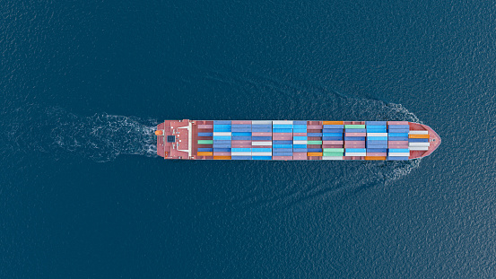 Aerial front view of a medium sized, loaded container cargo vessel traveling with speed over the ocean under cloudy sky