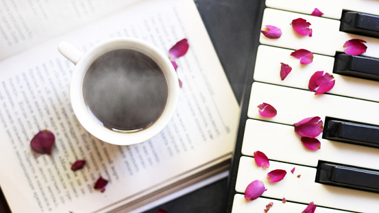 A cup of coffee on a book and a piano with rose petals, top view