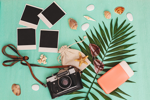 Summer background: palm leaf, seashells, camera and photos, sunscreen, and rose-colored sunglasses on a turquoise background