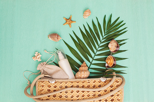 Wicker summer bag with beach accessories sunglasses, cosmetics and shells, with palm leaf on turquoise background, summer background