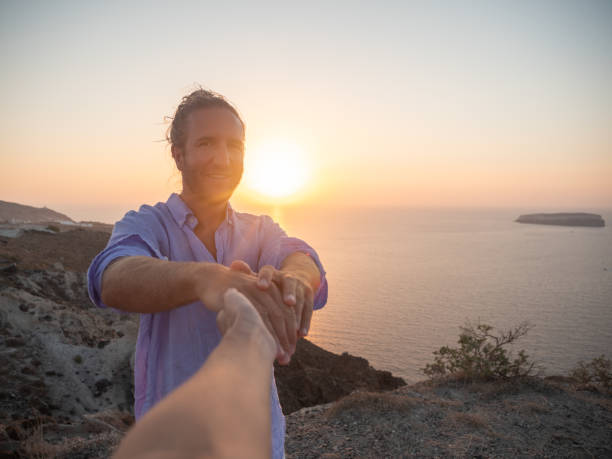 Couple holding hands on cliff at sunset Man leads the way happy couple on vacation in santorini greece stock pictures, royalty-free photos & images