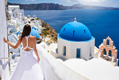 A beautiful tourist woman in a white summer dress looks at the blue domed church of the village of Oia, Santorini, Greece, during her summer holidays