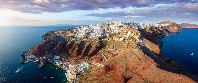 Panoramic aerial view to the village Oia on top of the caldera in Santorini, Greece, with the little fishing port Ammoudi just below during sunset time