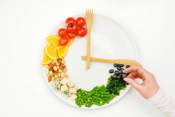 Colorful food and cutlery arranged in the form of a clock on a plate. Olive in hand. Intermittent fasting, diet, weight loss, lunch time concept. Colorful food and cutlery arranged in the form of a clock on a plate. Olive in hand. Intermittent fasting, diet, weight loss, lunch time concept. fork silverware table knife fine dining stock pictures, royalty-free photos & images