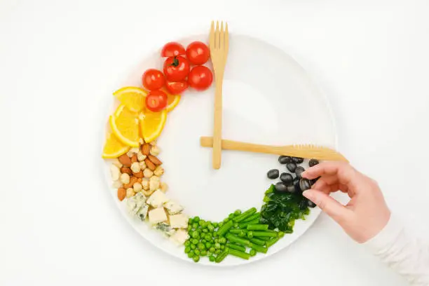 Colorful food and cutlery arranged in the form of a clock on a plate. Olive in hand. Intermittent fasting, diet, weight loss, lunch time concept.