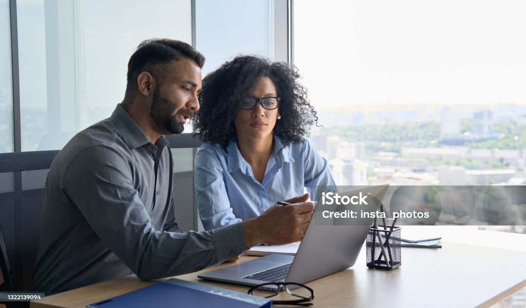 Multiethnic colleagues sitting at desk looking at laptop computer in office. Indian male ceo executive manager mentor giving consultation on financial operations to female African American colleague intern using laptop sitting in modern office near panoramic window. Advice Stock Photo